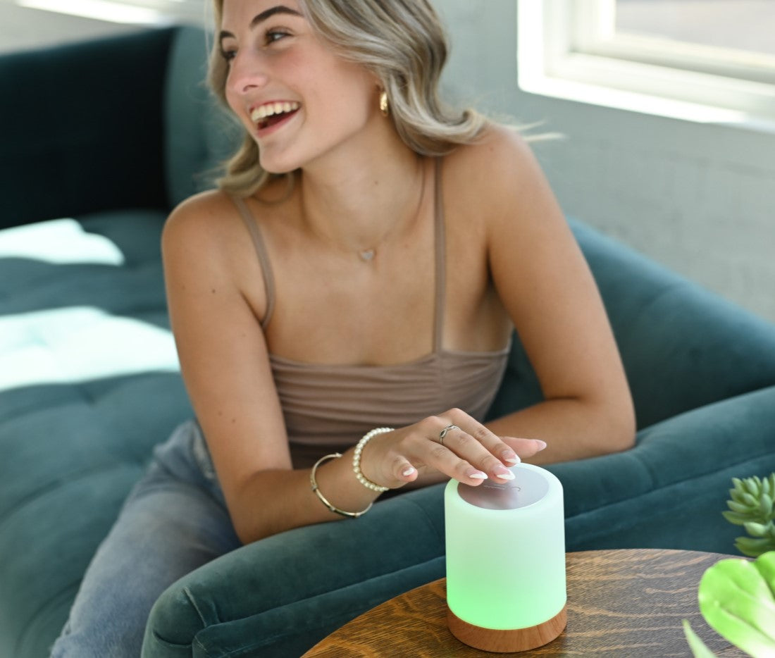 Woman smiling while touching a glowing green LuvLink Friendship Lamp on a wooden table in a modern living room.