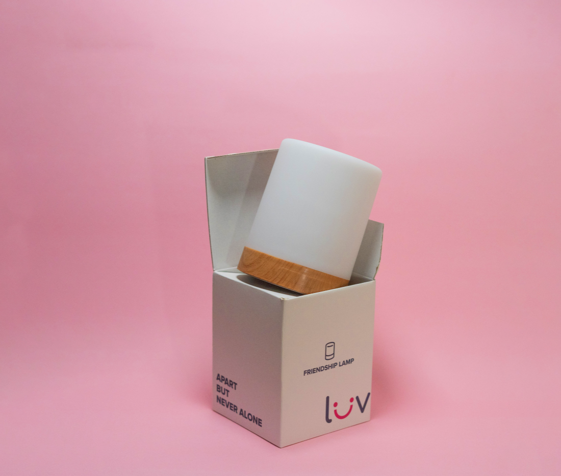 Luv Friendship Lamp with wooden base partially visible inside its white cardboard packaging against a pink background.